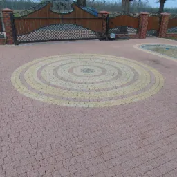 Ułożona kostka brukowa w okrągły wzór z dwóch kolorów, prowadząca do bramy wjazdowej z ceglanymi słupkami. Widoczny fragment trawnika.