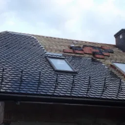 Układanie dachówki karpiówki na dachu z oknami połaciowymi w Skawie. Część dachu pokryta, część w trakcie prac. Widoczny komin i elementy konstrukcji.