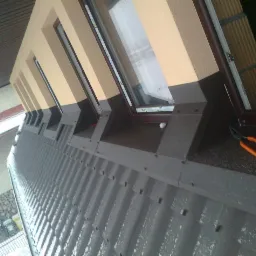 Szary dach z blachy trapezowej z oknami dachowymi i narzędziami. Widok z dołu na budynek w trakcie opadów śniegu.