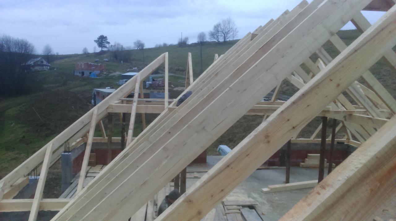 Konstrukcja drewniana więźby dachowej w trakcie montażu na tle pagórkowatego krajobrazu, widok z dołu, surowe drewno. Elementy muru w tle.