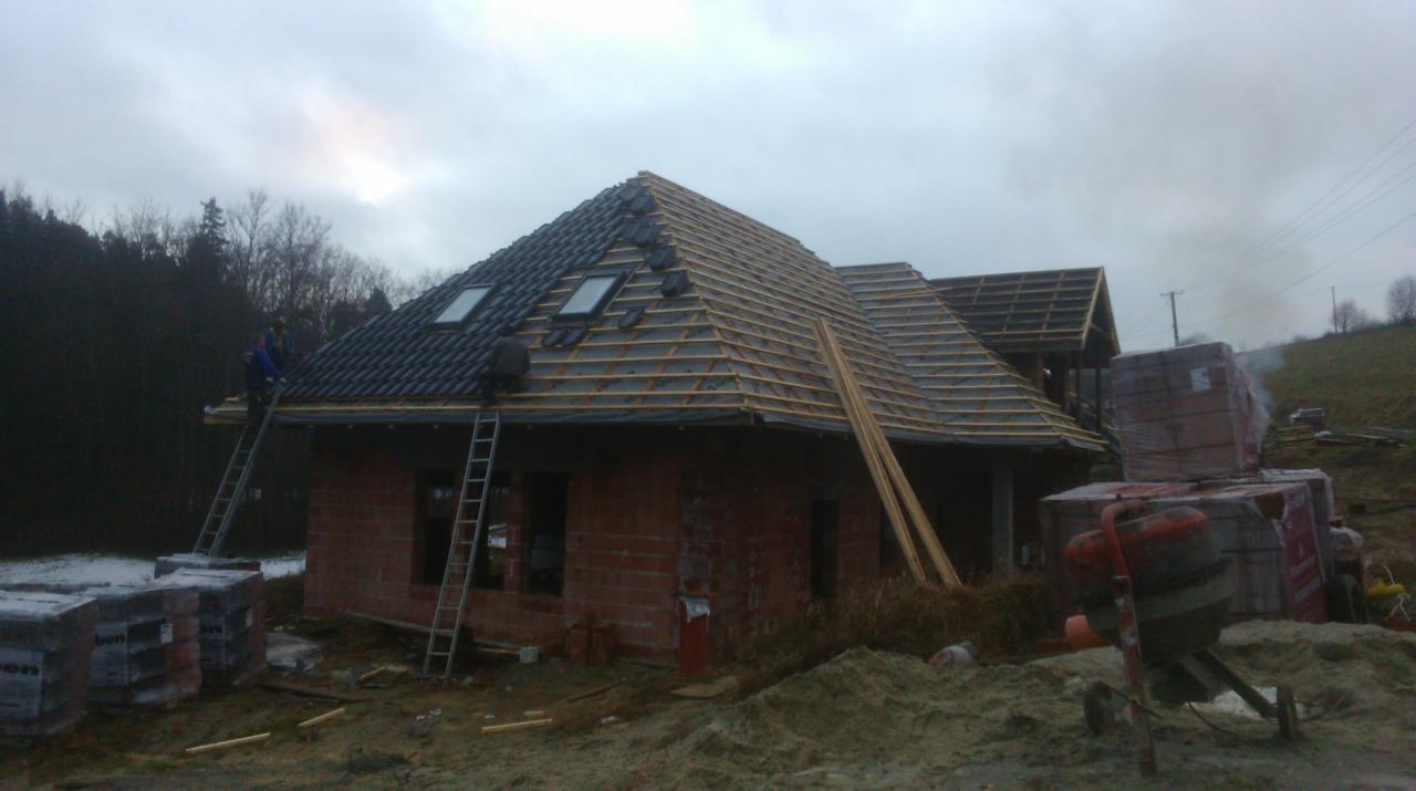 Montaż dachówki na domu jednorodzinnym; widoczne okna dachowe i drewniana konstrukcja. Pracownicy na drabinach układają pokrycie. W tle zalesione wzgórza, pochmurne niebo.