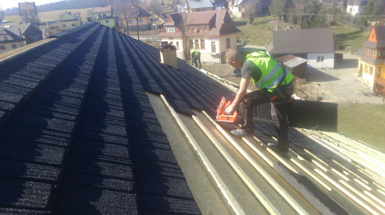 Praca na dachu: dekarz w kamizelce montuje łatę na przygotowanym dachu z ciemnej blachy. W tle zabudowa mieszkalna i wzgórza.
