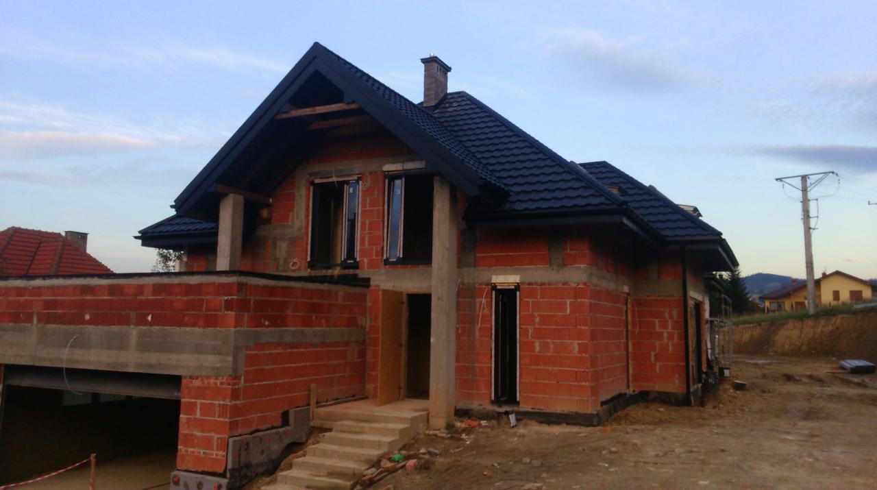 Niedokończony dom z czarnym dachem z dachówki ceramicznej, widoczne cegły i pustaki, okna bez szyb. Krajobraz wiejski w tle.