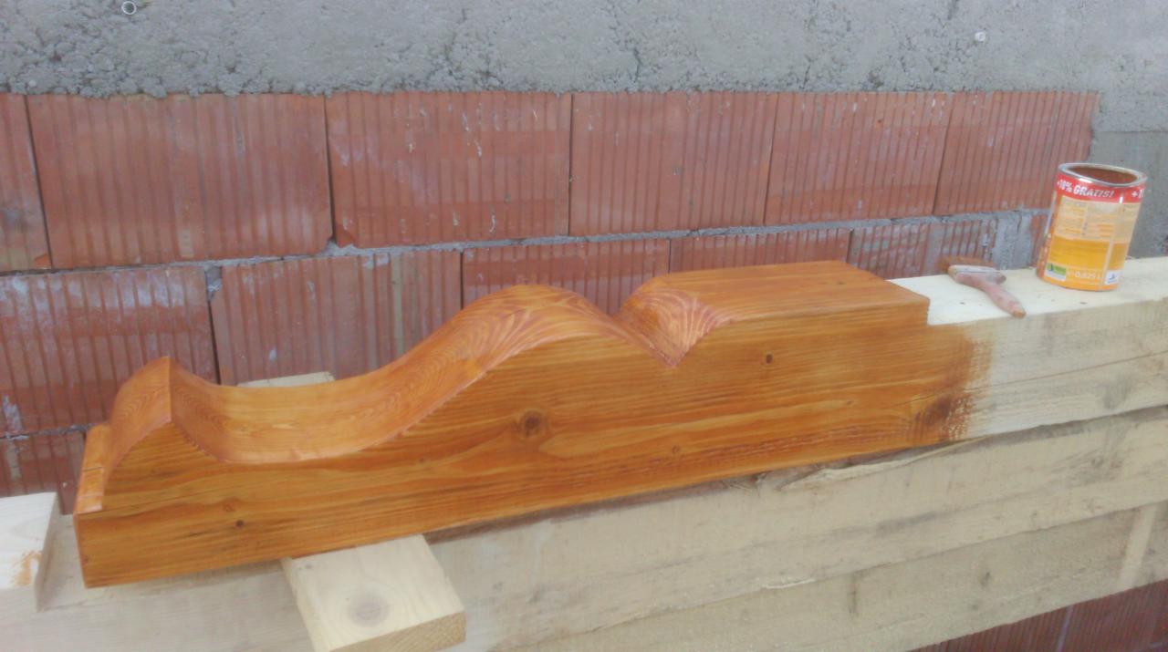 Impregnowana, dekoracyjna belka drewniana z falistym wykończeniem, częściowo pomalowana bejcą, leży na surowych deskach. W tle mur z cegły.