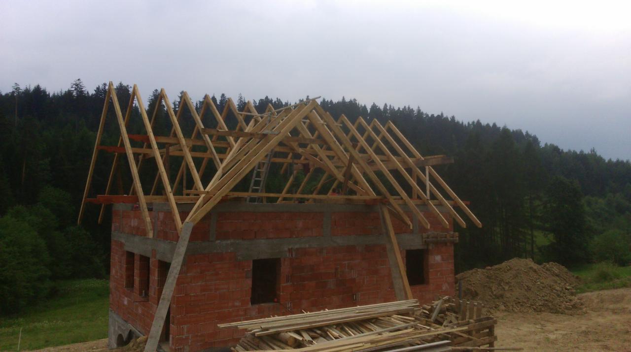Surowa więźba dachowa z jasnego drewna na murowanym budynku w trakcie budowy, na tle zalesionego wzgórza pod pochmurnym niebem.