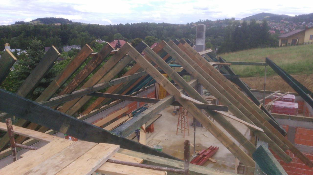 Wznoszenie konstrukcji dachu z drewnianych belek na murowanym budynku. Widoczne elementy więźby dachowej i materiały budowlane. W tle zalesione wzgórza.