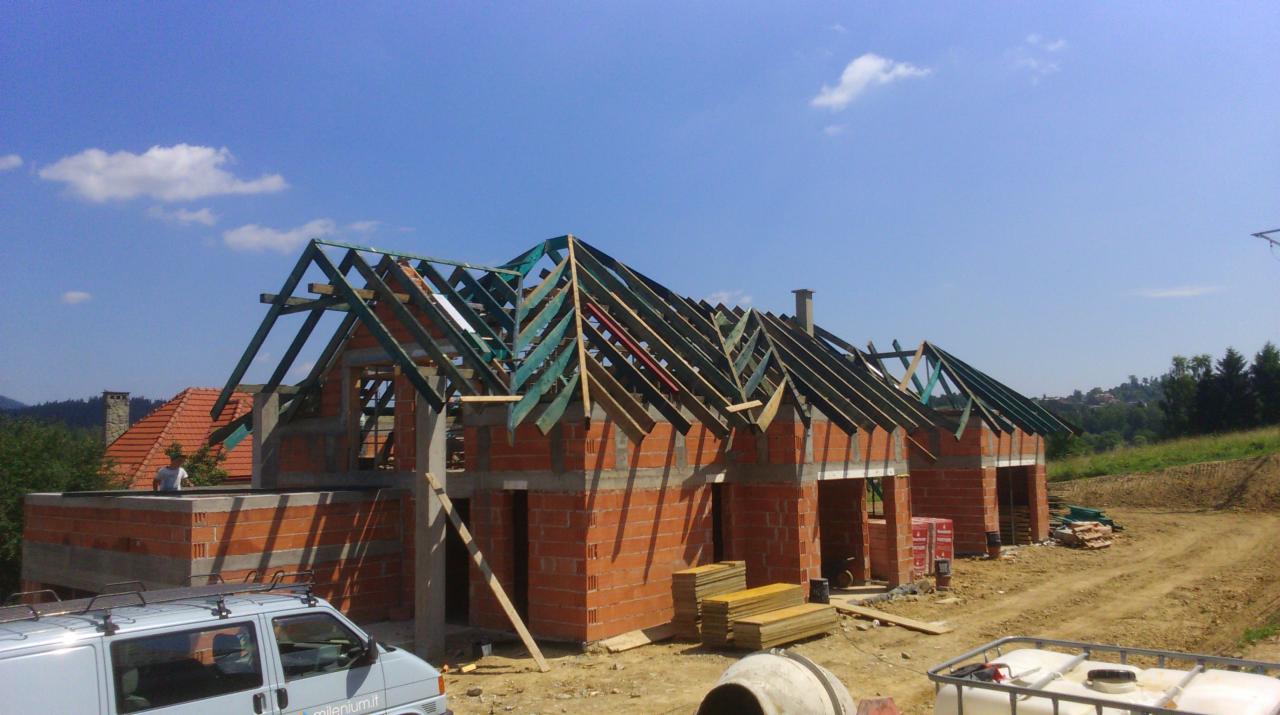 Surowa konstrukcja dachu z więźbą na murowanym budynku w trakcie budowy, widoczna z ziemi. W tle zieleń i błękit nieba.