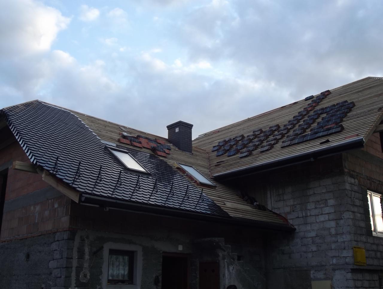 Montaż dachówki karpiówki na połaci dachu domu jednorodzinnego z oknami dachowymi. Część dachu pokryta, reszta w trakcie prac. Widoczny komin i fragment elewacji budynku.