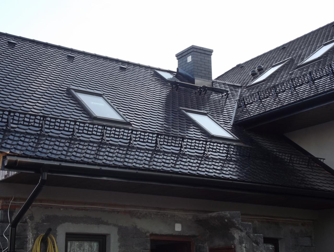 Czarny dach z dachówką karpiówką, trzema oknami dachowymi i kominem. Rynna wzdłuż krawędzi dachu. Widok na fragment elewacji budynku.