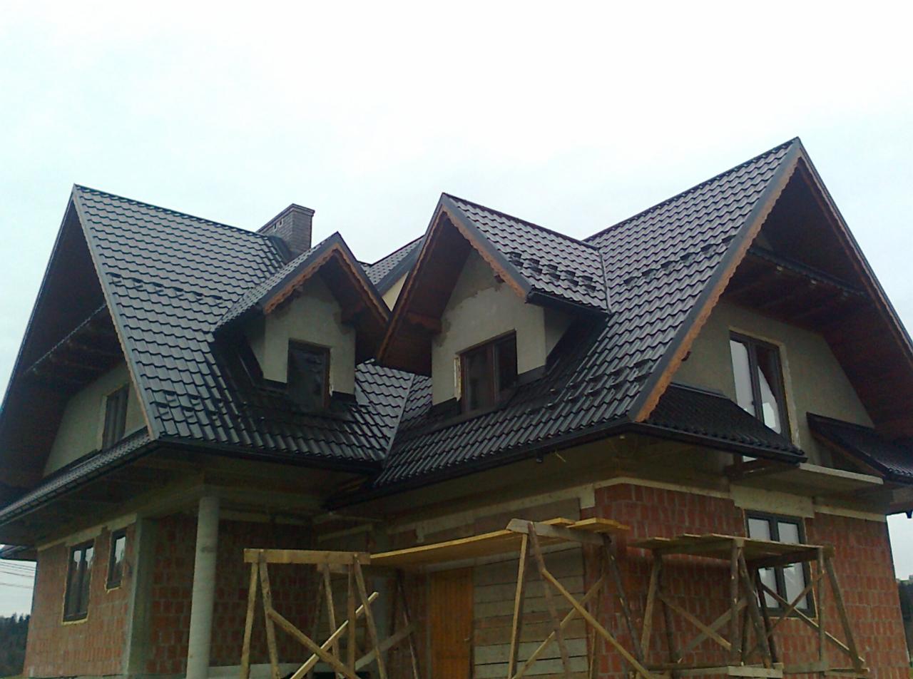 Dom w trakcie budowy z czarną dachówką karpiówką. Widoczne rusztowania i fragmenty muru z cegły. Dach z lukarnami. Pochmurne niebo w tle.