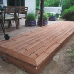 Nowy, brązowy taras drewniany z desek, z widocznym grillem i meblami ogrodowymi w tle. Taras otoczony zielenią.