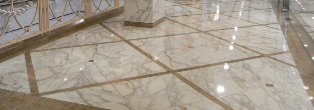 Błyszcząca podłoga z białego marmuru z brązowymi intarsjami tworzącymi geometryczny wzór, widoczna fragment poręczy z dekoracyjnymi elementami.