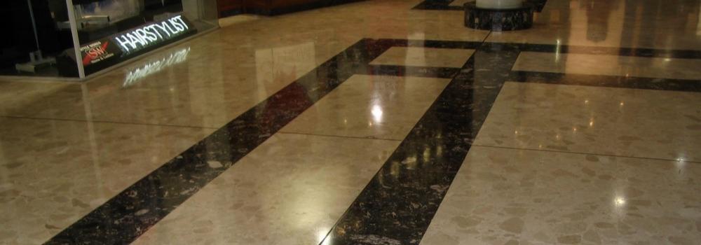 Polerowana marmurowa posadzka w holu z ciemnymi, żyłkowatymi wstawkami układającymi się w geometryczny wzór, odbijająca światło i widok witryny salonu fryzjerskiego.