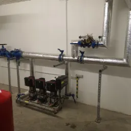 Instalacja grzewcza z izolowanymi rurami, zaworami i czterema pompami obiegowymi Grundfos w kotłowni z czerwonym zbiornikiem wyrównawczym.