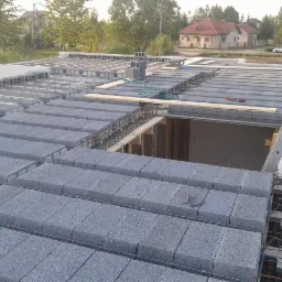 Wznoszenie stropu Teriva z szarych bloczków i żebrowanych belek, widoczne zbrojenie, drabina oparta o krawędź stropu, w tle zabudowa jednorodzinna.