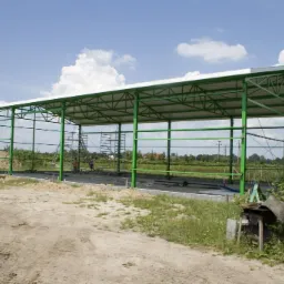 Konstrukcja stalowa z zielonymi słupami i białym dachem w trakcie montażu na tle błękitnego nieba z chmurami i krajobrazu wiejskiego. Widoczne rusztowania.