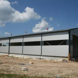 Hala z płyty warstwowej w trakcie montażu, widoczne niezabudowane otwory okienne i drzwiowe, czerwone elementy na dachu, teren budowy.