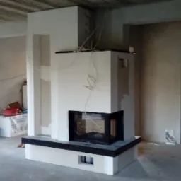 Wnętrze w trakcie prac wykończeniowych: surowy betonowy strop, ściany po nałożeniu gładzi, w centrum biały kominek z czarnymi elementami wykończeniowymi, widoczne przewody elektryczne...