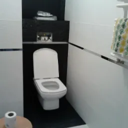 Biała toaleta w niszy z czarnymi płytkami, uzupełniona o półkę z akcesoriami. Jasne, kafelkowane ściany z metalową listwą i ręcznik na grzejniku.