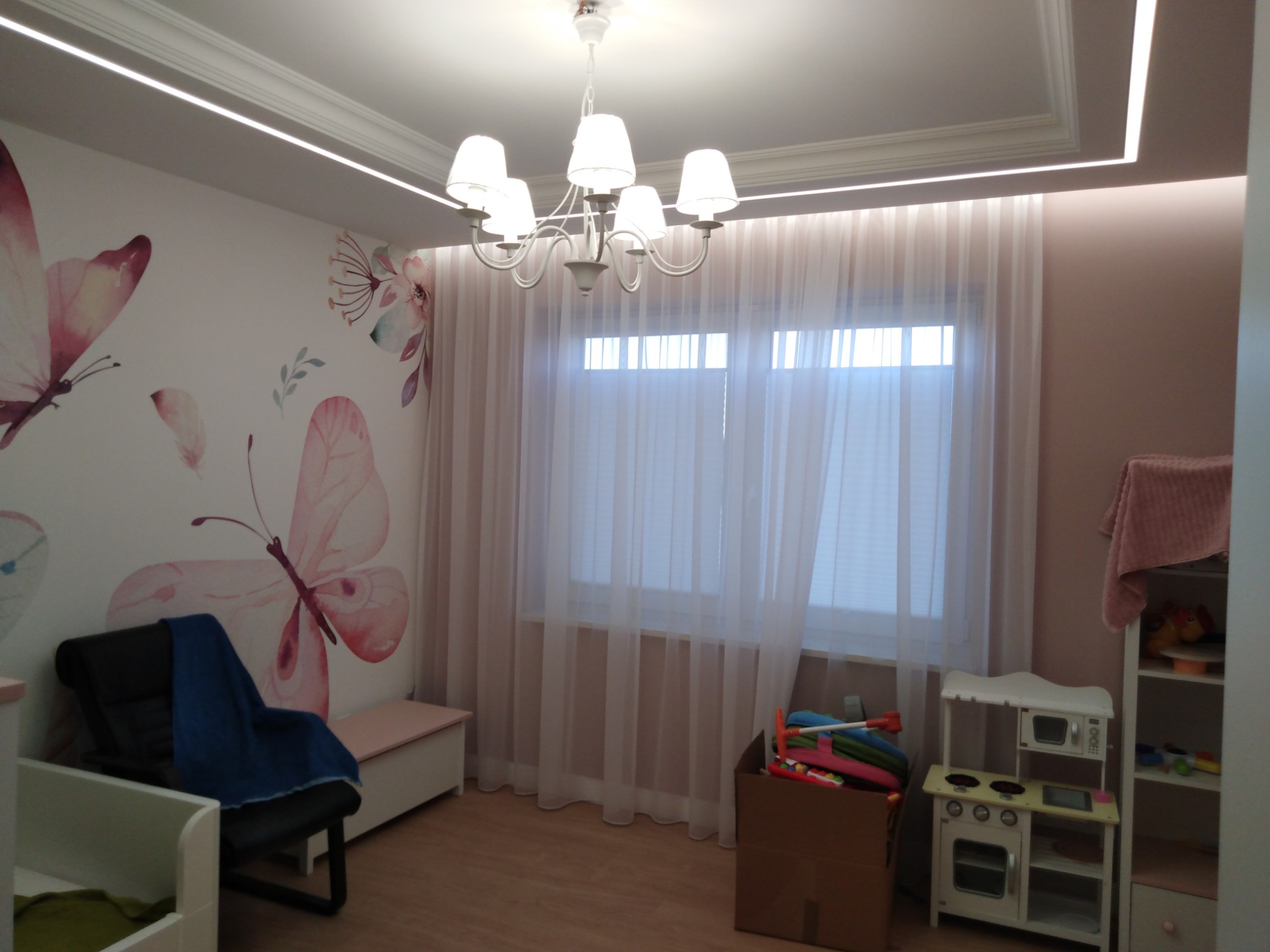 Pokój dziecięcy z motywem motyli na ścianie, oświetleniem LED w suficie podwieszanym i białym żyrandolem. W pokoju znajduje się mała kuchnia dla dzieci i karton z zabawkami.