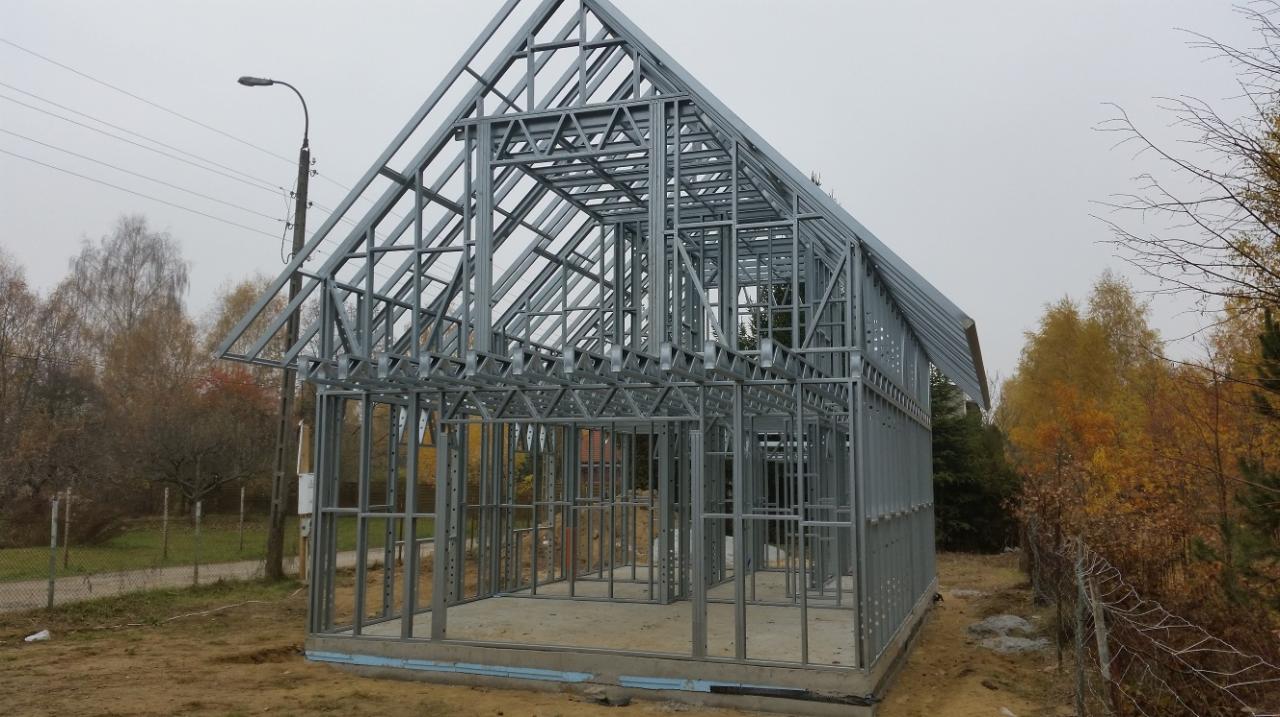 Konstrukcja szkieletowa domu w trakcie budowy, widoczne stalowe profile, jesienne drzewa w tle i słup oświetleniowy. Fundament z izolacją.