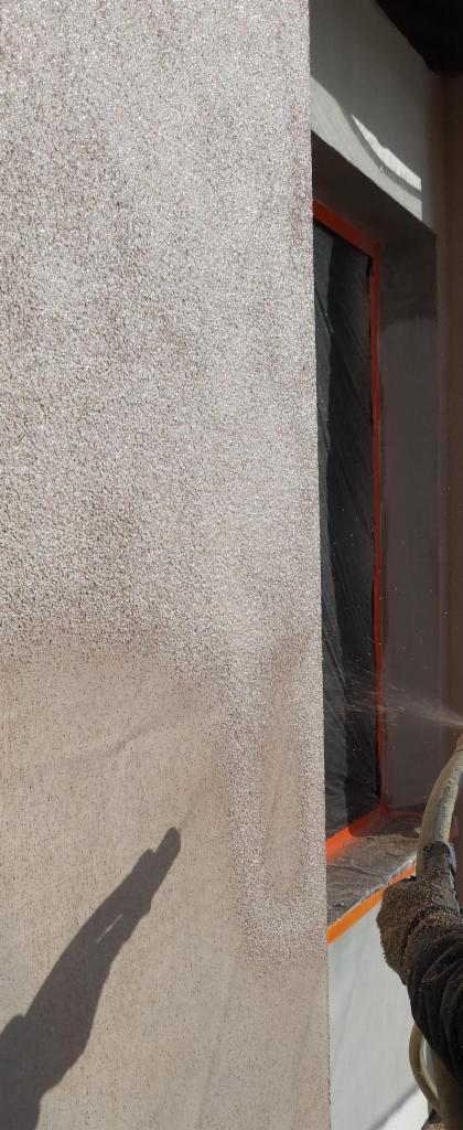 Nakładanie struktury elewacyjnej metodą natryskową na ścianę budynku, okno zabezpieczone folią i taśmą malarską.