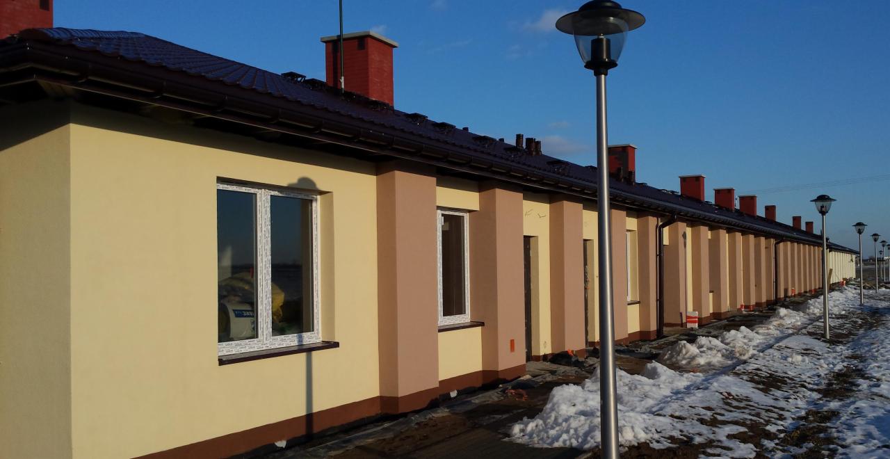 Długi szereg nowo wybudowanych, parterowych budynków w kolorze beżowym i brązowym, z czerwonymi kominami i oknami z PCV, oświetlonych przez latarnie uliczne wzdłuż zasypanej śniegiem drogi.