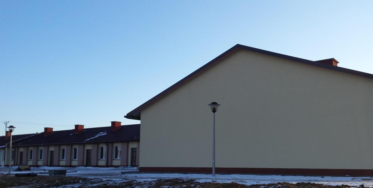 Nowe budynki mieszkalne w zabudowie szeregowej, wykończone elewacją w kolorze beżowym, z brązowymi dachami i kominami. Na pierwszym planie lampa oświetleniowa i resztki śniegu.