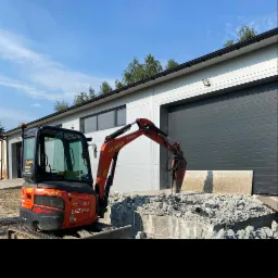 Pomarańczowa minikoparka Kubota z młotem hydraulicznym rozbija betonową konstrukcję na tle szarego budynku z bramami rolowanymi i błękitnego nieba.