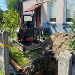 Mała koparka OMAX stoi obok wykopu pod fundamenty domu jednorodzinnego, obok leży rura kanalizacyjna i butelka wody.