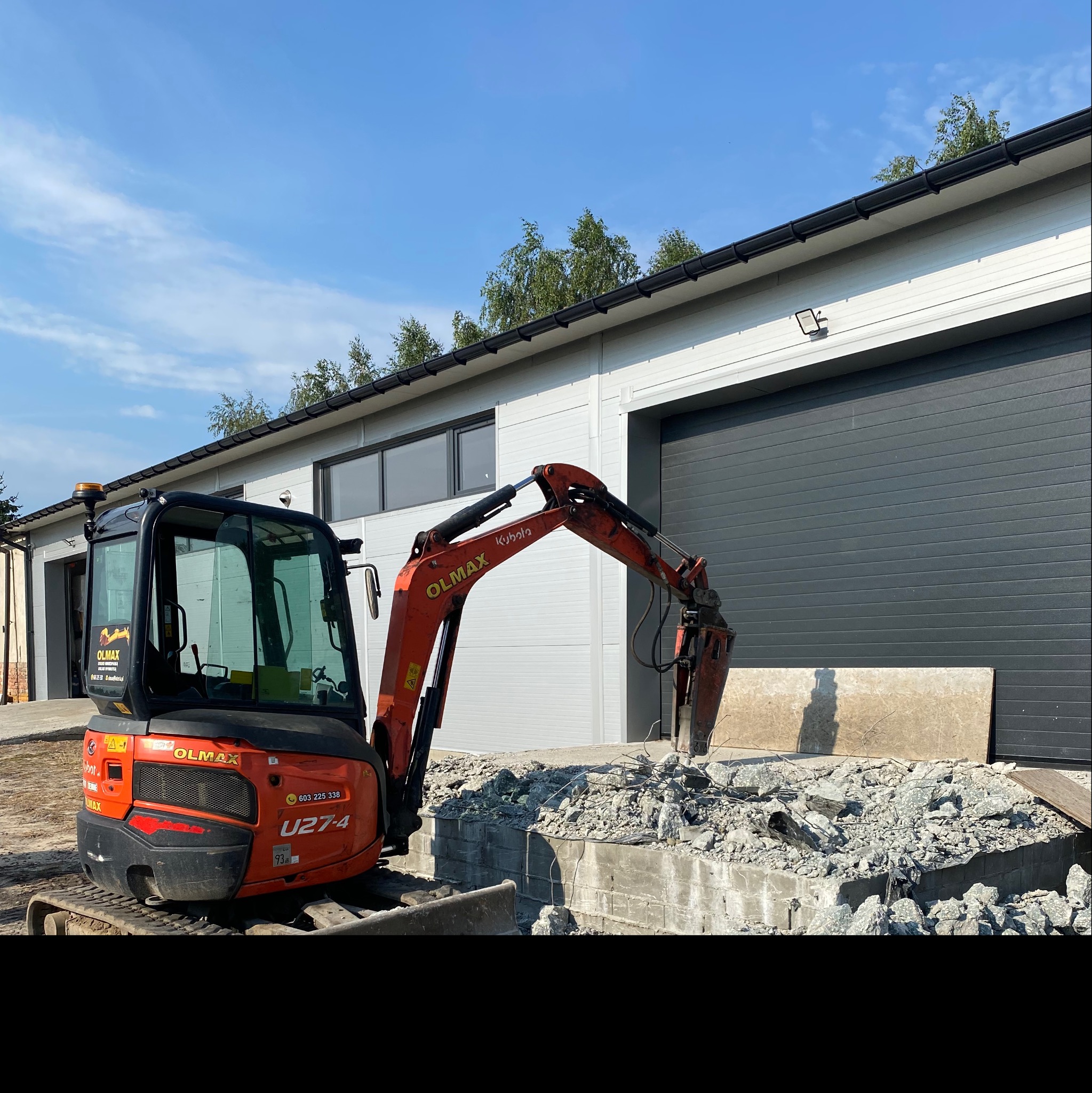 Pomarańczowa minikoparka Kubota z młotem hydraulicznym rozbija betonową konstrukcję na tle szarego budynku z bramami rolowanymi i błękitnego nieba.