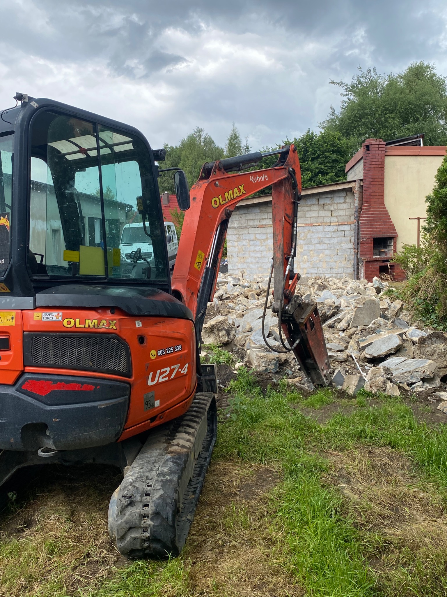 Pomarańczowa mini koparka Kubota z hydraulicznym młotem na tle częściowo zburzonego budynku z czerwonej cegły i gruzu, pochmurne niebo.