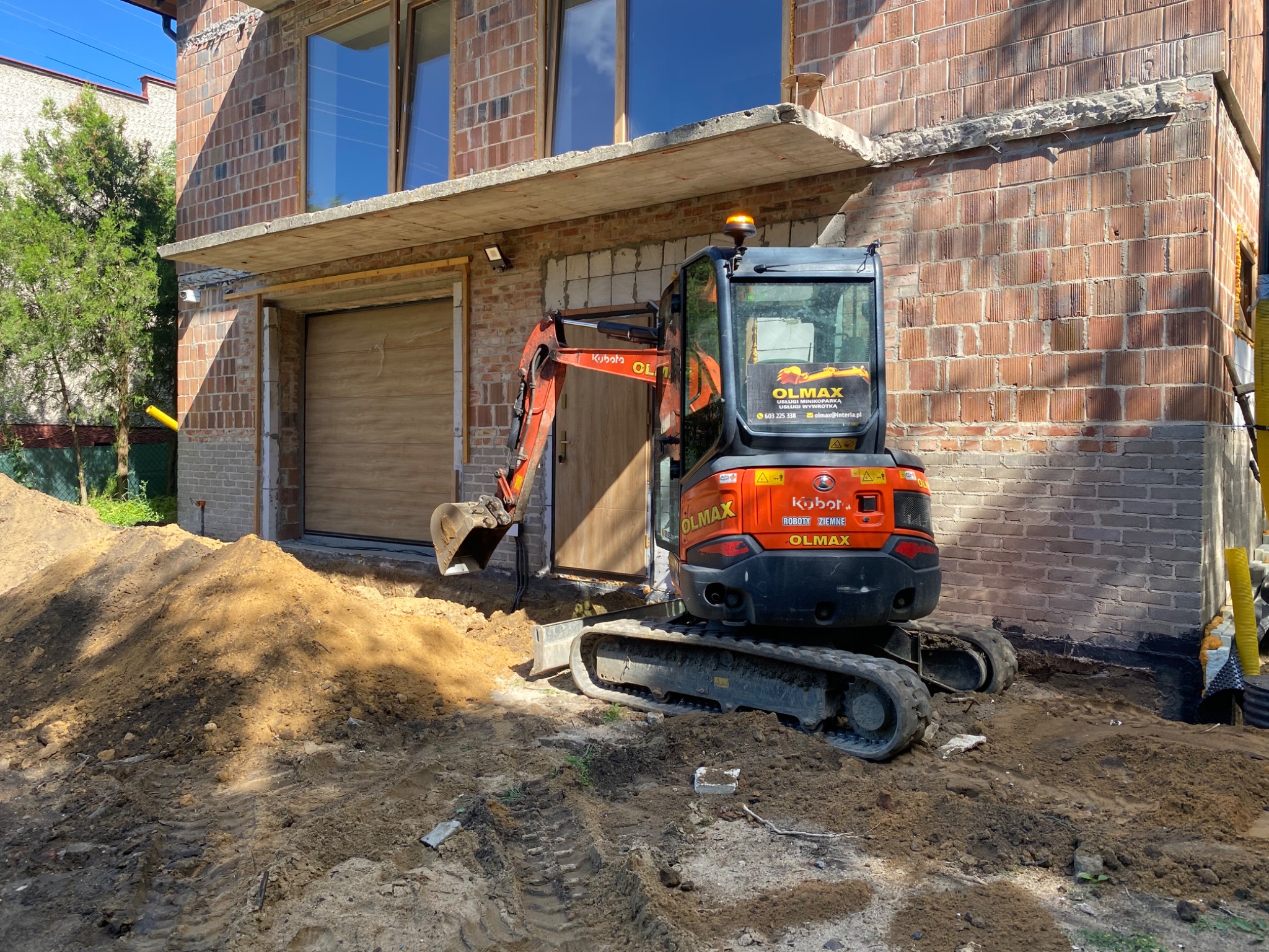 Pomarańczowa minikoparka Kubota marki Olmax na placu budowy, z wykopaną ziemią przed budynkiem z cegły, częściowo otynkowanym, z widocznymi oknami i bramą garażową.