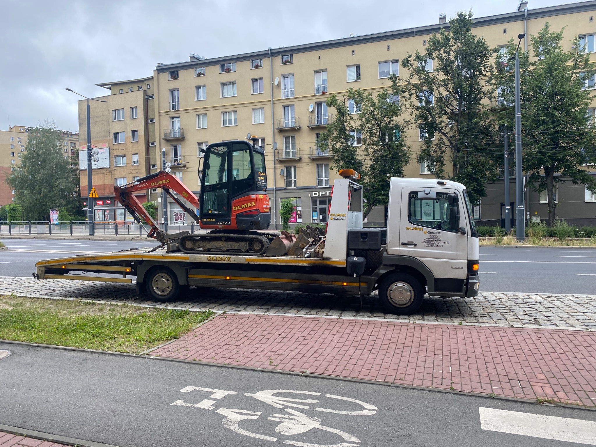 Laweta z minikoparką Kubota U27-4 na tle miejskiej zabudowy, widoczny znak ścieżki rowerowej na pierwszym planie i reklama firmy Olmax na lawetce.