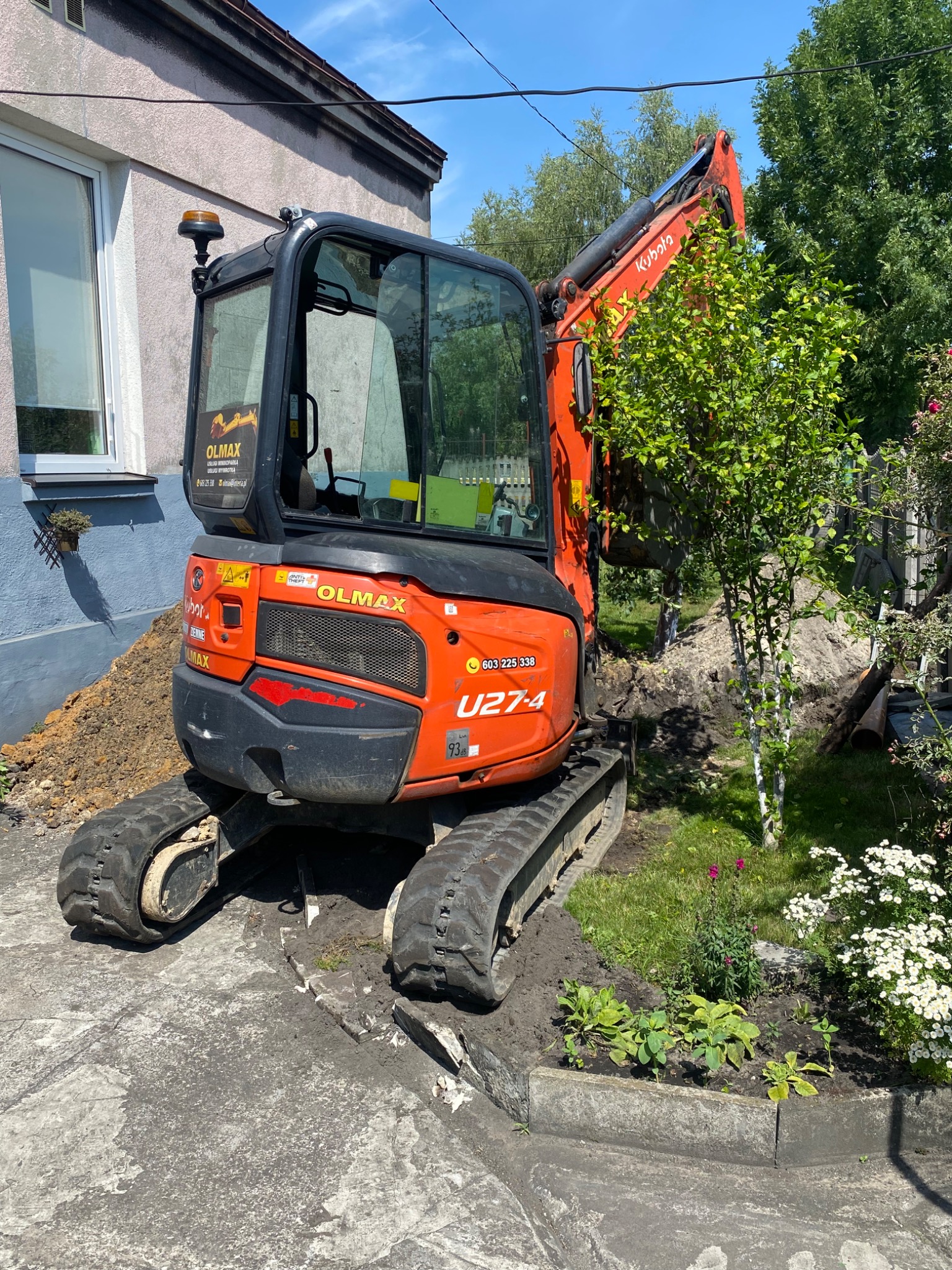 Pomarańczowo-czarna mini koparka Kubota U27-4 firmy Olmax na betonowej nawierzchni, obok wykopana ziemia i zieleń, w tle budynek mieszkalny.