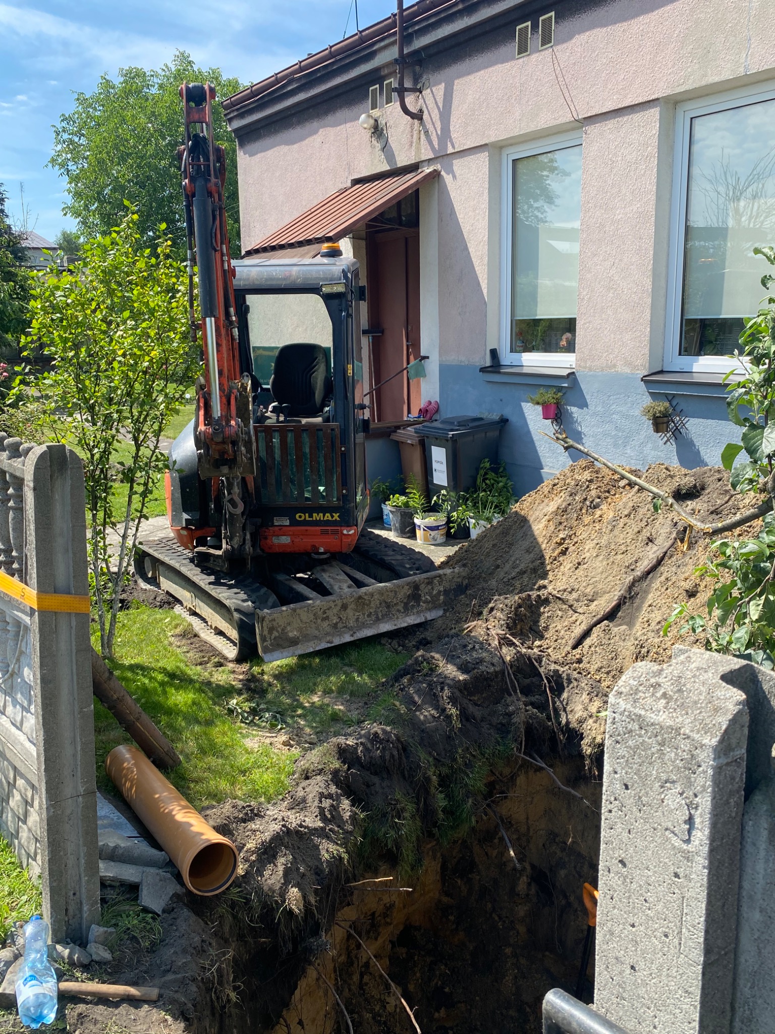 Mała koparka OMAX stoi obok wykopu pod fundamenty domu jednorodzinnego, obok leży rura kanalizacyjna i butelka wody.