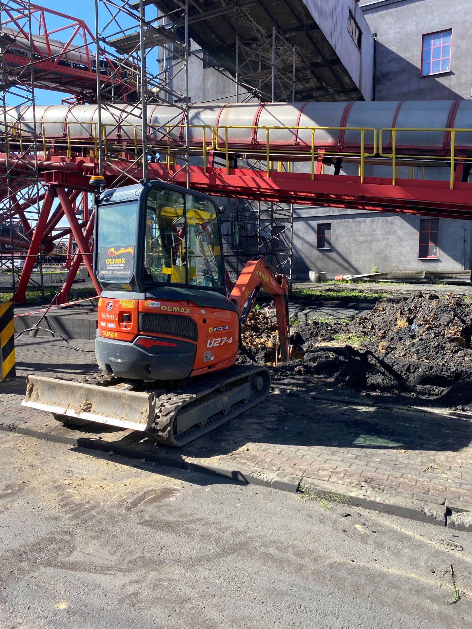 Pomarańczowa mini koparka Kubota U27-4 marki Olmax na placu budowy z widocznym stosem ziemi i konstrukcją taśmociągu w tle.