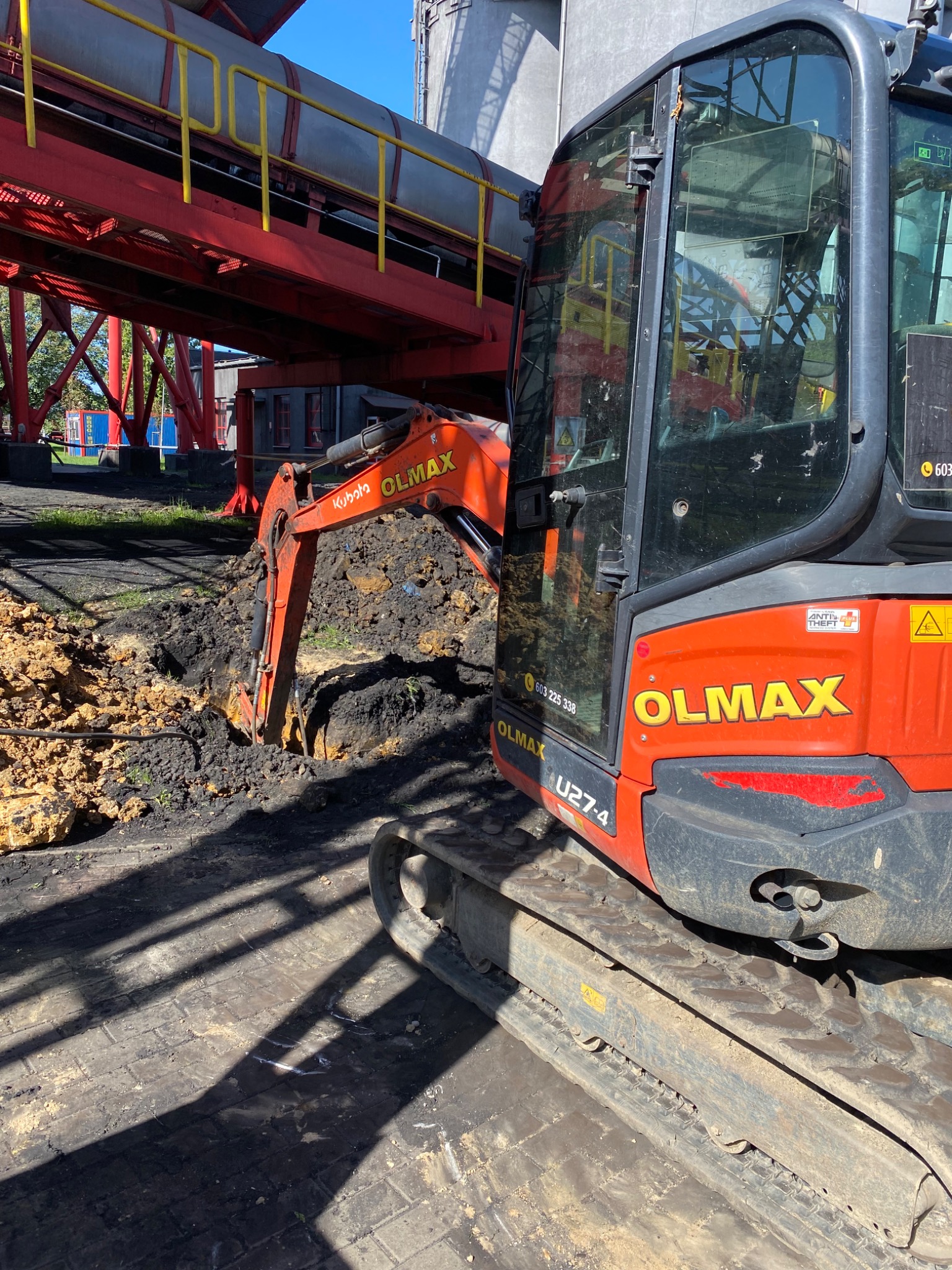 Pomarańczowa mini koparka Kubota OLMAX U27-4 wykonuje wykop w ziemi, w tle konstrukcja przemysłowa z rurociągami.