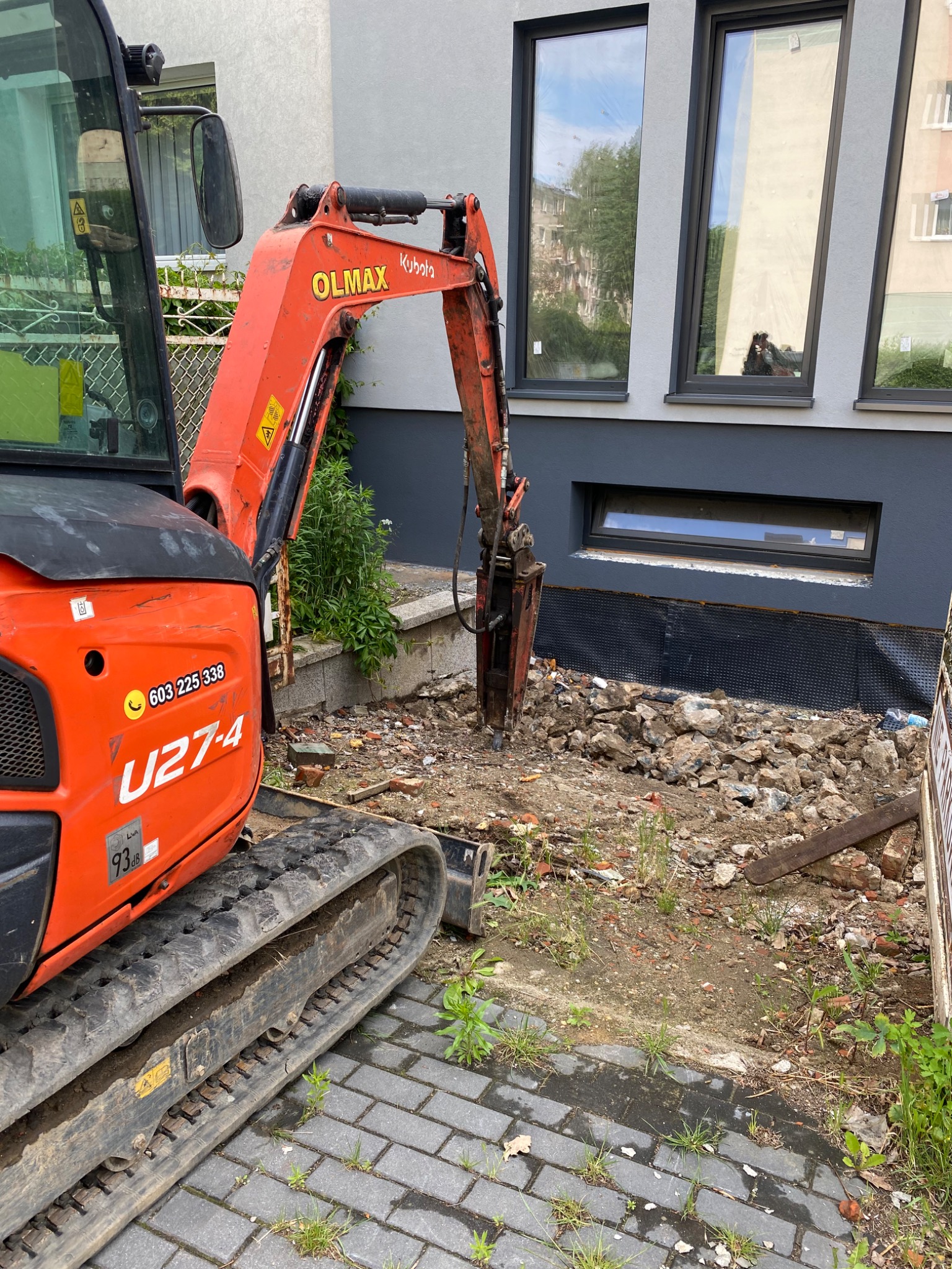 Pomarańczowa minikoparka Kubota z młotem hydraulicznym w trakcie prac przy budynku, gruz i ziemia w wykopie, widoczne okna budynku. Na korpusie koparki naklejka z numerem telefonu.