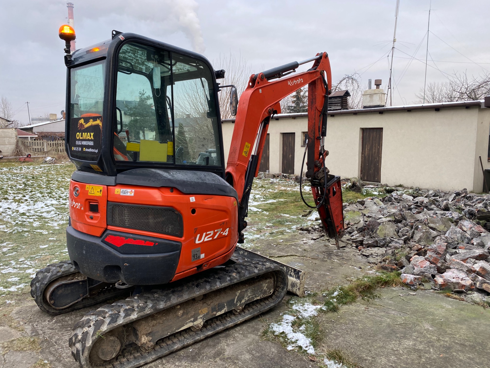 Pomarańczowa minikoparka Kubota U27-4 z hydraulicznym młotem na tle sterty gruzu i budynku gospodarczego w pochmurny dzień.
