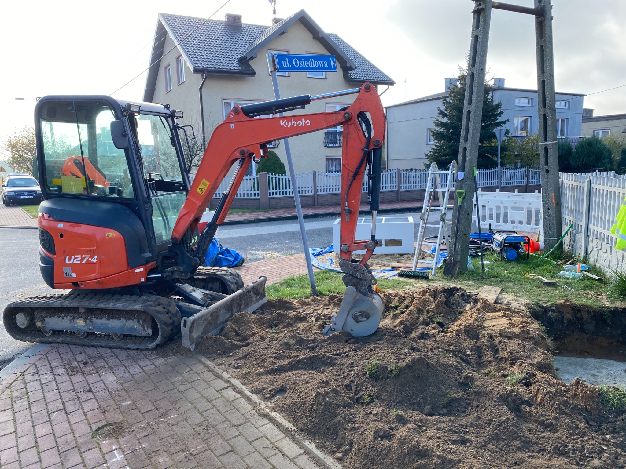 Pomarańczowa mini koparka Kubota U27-4 na kostce brukowej obok wykopu w ziemi, w tle dom z tabliczką 'ul. Osiedlowa' i słupy energetyczne.