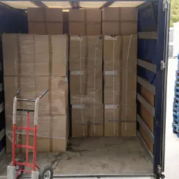 Wnętrze otwartej naczepy z kartonami zabezpieczonymi folią stretch, czerwony wózek transportowy i stosy drewnianych palet obok.