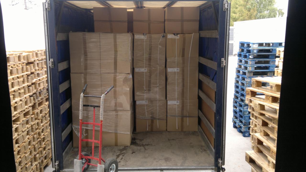 Wnętrze otwartej naczepy z kartonami zabezpieczonymi folią stretch, czerwony wózek transportowy i stosy drewnianych palet obok.