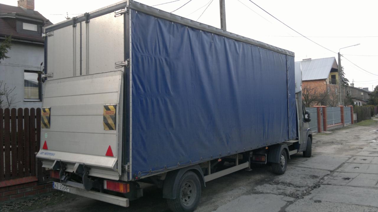 Ciężarówka z plandeką w kolorze granatowym, zaparkowana na ulicy. Widoczny tył pojazdu z otwartymi drzwiami. W tle zabudowa jednorodzinna.