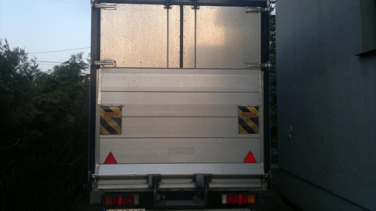 Zamknięte tylne drzwi aluminiowej naczepy ciężarówki z widocznymi elementami ostrzegawczymi i światłami. Widoczne drzewa i budynek w tle.