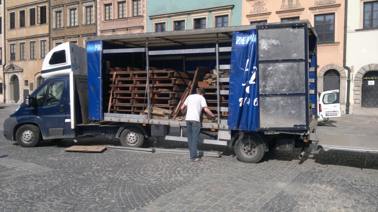 Niebieski bus dostawczy z otwartą przestrzenią ładunkową, wypełnioną paletami i elementami drewnianymi. Mężczyzna w białej koszulce przy pojeździe na brukowanym placu miejskim.