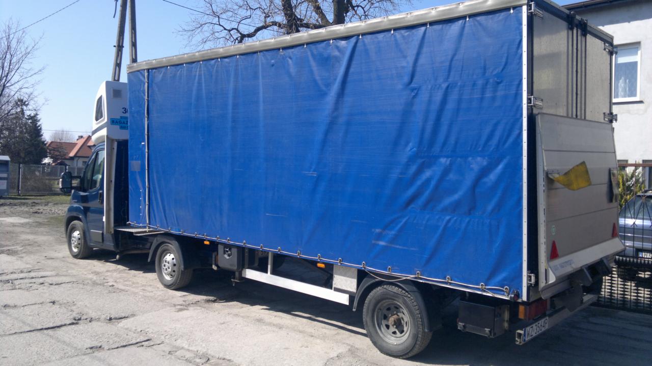 Ciężarówka z plandeką, zaparkowana na szarym asfalcie. Widoczny bok pojazdu z niebieską plandeką i fragment budynku w tle.