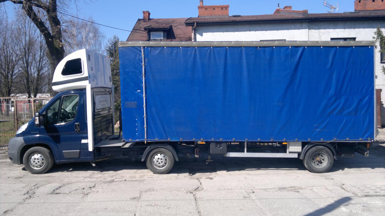 Niebieski bus dostawczy z plandeką, widok z boku na tle miejskiej zabudowy. Charakterystyczna nadbudówka kabiny sypialnej.