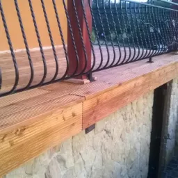 Drewniany taras z kutą balustradą i kamienną elewacją. Widok z boku, podkreślający detale łączenia desek i solidność konstrukcji. W tle zieleń ogrodu i fragment zaparkowanego auta.