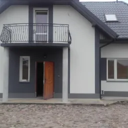 Dwukondygnacyjny dom z balkonem, pomalowany na biało-szaro, z otwartymi drzwiami wejściowymi i widocznym fragmentem elewacji wykończonej na gładko, na pierwszym planie kostka brukowa i składowane...